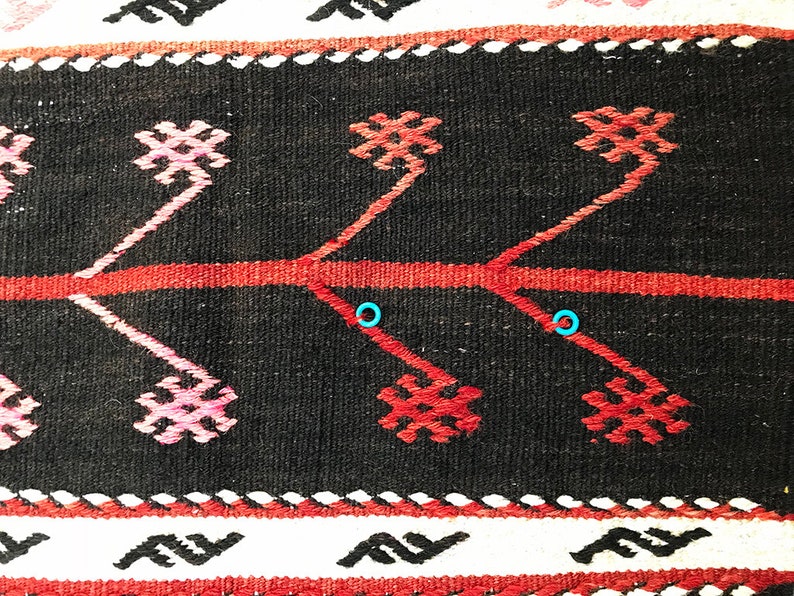 May include: Close-up of a black and red woven rug with a geometric pattern. The rug has a white border with a repeating pattern of stylised waves.