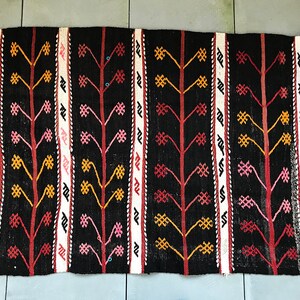 May include: A black and white rug with a repeating pattern of stylised trees with red, pink, and yellow accents. The rug has a fringe border.