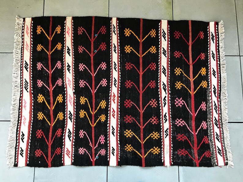 May include: A black and white rug with a repeating pattern of stylised trees with red, pink, and yellow accents. The rug has a fringe border.