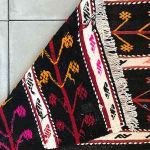 May include: A black, white, red and yellow patterned rug with fringe. The rug has a geometric design with a repeating pattern of stylised flowers and leaves.