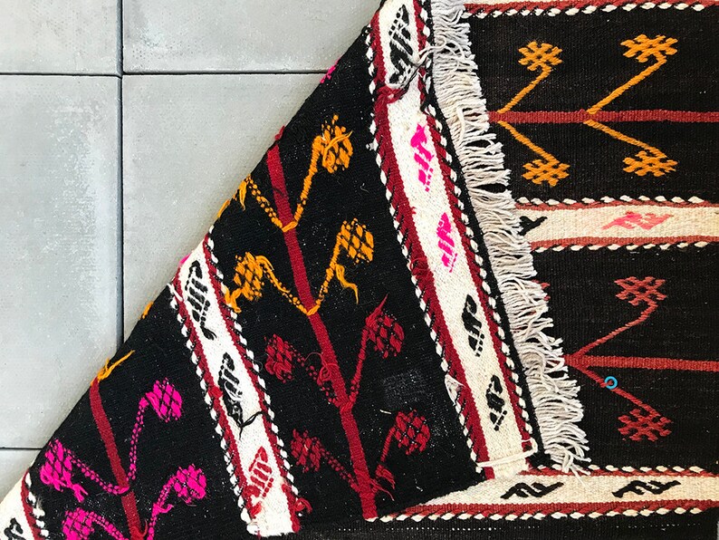 May include: A black, white, red and yellow patterned rug with fringe. The rug has a geometric design with a repeating pattern of stylised flowers and leaves.
