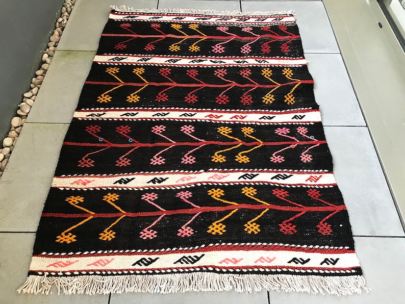 May include: A black and white rug with a geometric pattern in red, orange, and pink. The rug has a fringe border.