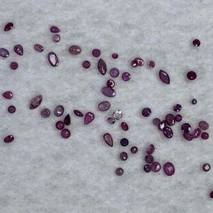May include: A collection of loose, polished gemstones in various shapes and sizes, including round, oval, and marquise cuts. The gems are primarily shades of deep red and purple, scattered on a white, textured surface. One gem appears to be silver.