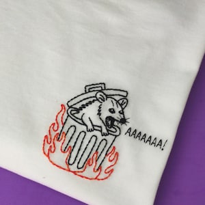 May include: A white t-shirt featuring an embroidered design of an opossum inside a trash can surrounded by orange flames. The text AAAAAAA! is embroidered in black thread to the right of the trash can, completing the graphic illustration.
