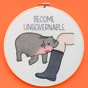 Puede incluir: Arte de bordado en aro con un hipopótamo de dibujos animados mordiendo una pierna humana con el texto "Become Ungovernable".
