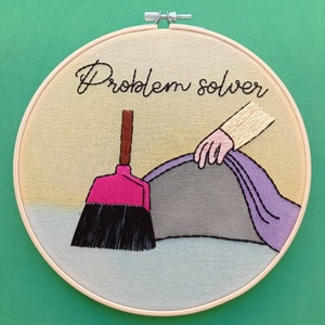 Può includere: Un telaio da ricamo circolare contenente un pannello in tessuto beige con il testo ricamato in nero Problem solver. Il disegno mostra una mano che solleva un panno viola per rivelare una superficie grigia, con una scopa rosa e nera.
