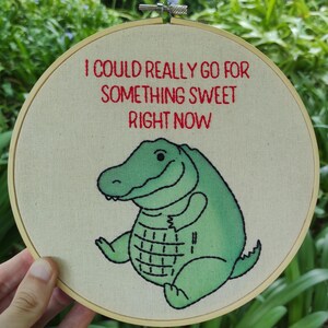 May include: Embroidered hoop featuring the text "I COULD REALLY GO FOR SOMETHING SWEET RIGHT NOW" in red. A green alligator illustration is below the text. The alligator has a smiling face and a grid pattern on its belly.
