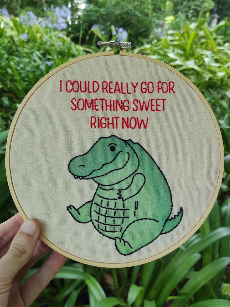 May include: Embroidered hoop featuring the text "I COULD REALLY GO FOR SOMETHING SWEET RIGHT NOW" in red. A green alligator illustration is below the text. The alligator has a smiling face and a grid pattern on its belly.
