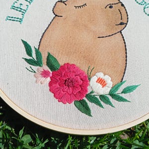 May include: Embroidered artwork featuring a capybara with closed eyes, surrounded by a floral arrangement. The capybara is tan, outlined in black, and the flowers are pink, white, and green. The text "LE" is visible in green.