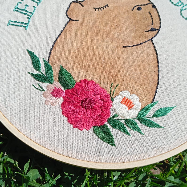 May include: Embroidered artwork featuring a capybara with closed eyes, surrounded by a floral arrangement. The capybara is tan, outlined in black, and the flowers are pink, white, and green. The text "LE" is visible in green.