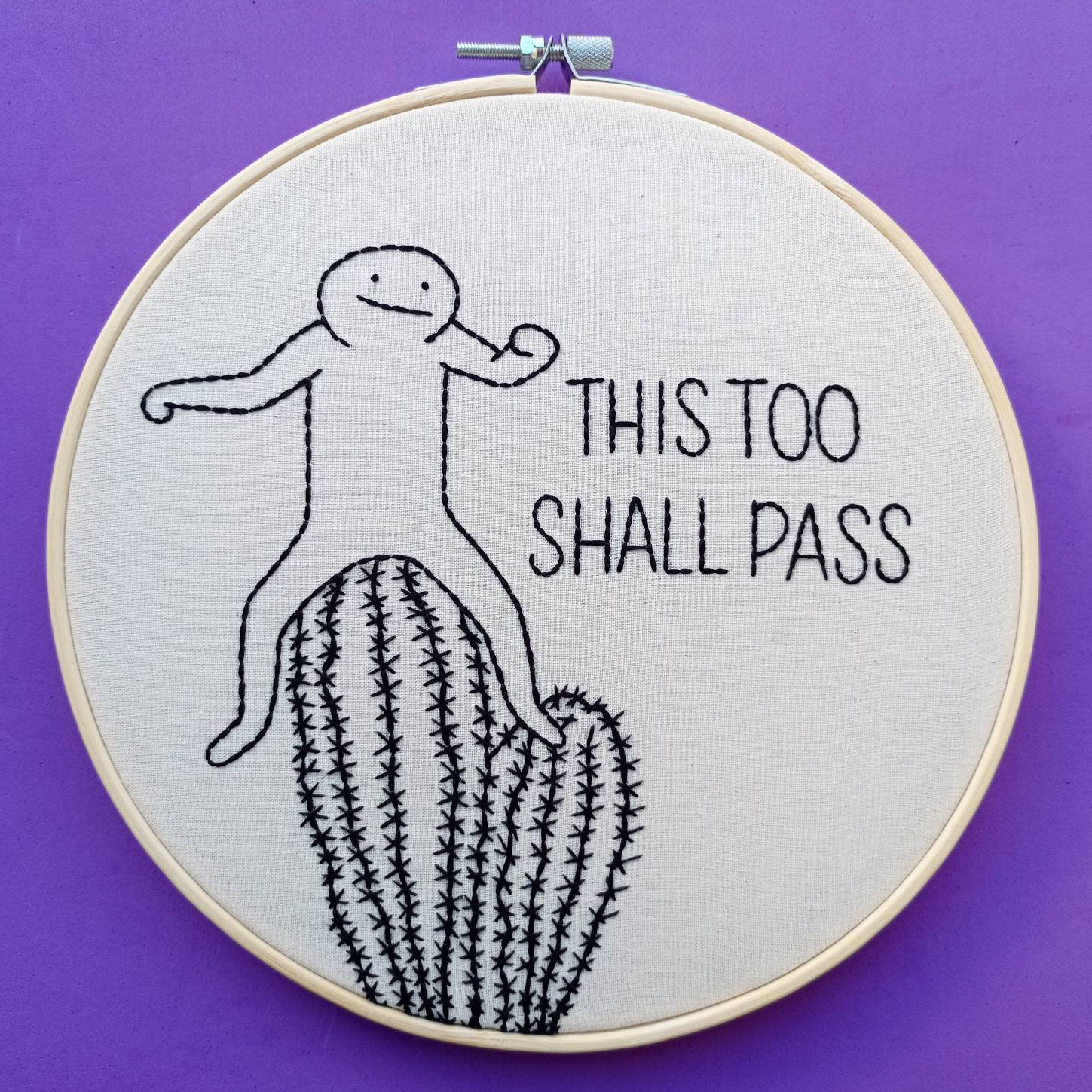 May include: Embroidered hoop art with a stick figure standing on a cactus. The text "This too shall pass" is embroidered below the figure.