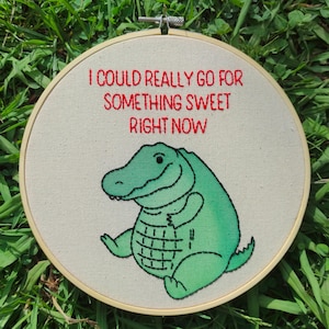 May include: Embroidered hoop art with a green alligator and the red text: "I COULD REALLY GO FOR SOMETHING SWEET RIGHT NOW". The alligator is smiling. The background is a light beige fabric, set in a wooden hoop.
