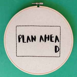 May include: A white embroidery hoop with a black embroidered rectangle that says "PLAN AHEAD" with a "D" at the bottom right corner.