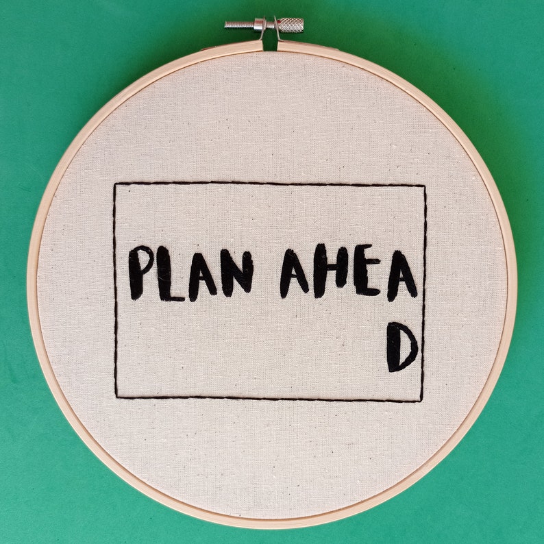 May include: A white embroidery hoop with a black embroidered rectangle that says "PLAN AHEAD" with a "D" at the bottom right corner.