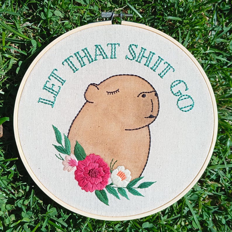 May include: Embroidered hoop art featuring a capybara with the text "LET THAT SHIT GO" in green thread. The capybara is tan with closed eyes, surrounded by embroidered flowers in pink, white, and green. The hoop is made of wood.