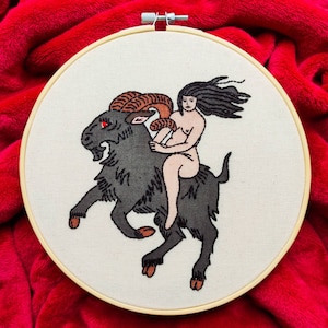 Puede incluir: Arte de bordado en aro que representa a una mujer montando una cabra negra con cuernos marrones. La mujer tiene el pelo largo y negro y está desnuda. El fondo es una tela blanca.
