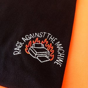 Può includere: Maglietta nera con ricamo bianco di una stampante in fiamme e il testo "Rage Against The Machine" intorno.