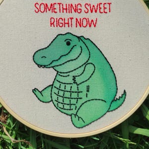May include: Embroidered hoop art showing a smiling green alligator with black outlines. The text "SOMETHING SWEET RIGHT NOW" is stitched in red above the alligator. The artwork is set against a cream-coloured fabric.