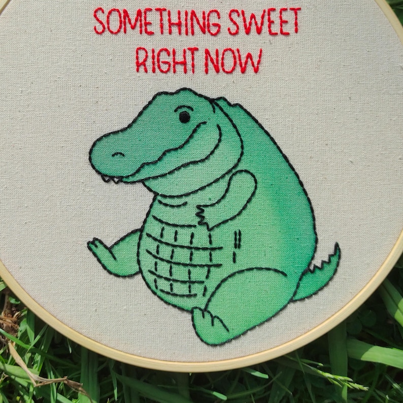 May include: Embroidered hoop art showing a smiling green alligator with black outlines. The text "SOMETHING SWEET RIGHT NOW" is stitched in red above the alligator. The artwork is set against a cream-coloured fabric.