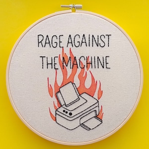 May include: Embroidered hoop art featuring a printer on fire with the text "Rage Against The Machine" in black lettering.