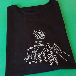 May include: Black t-shirt with white embroidered design of a dinosaur looking up at a falling comet labeled "The Economy". The text "Oh no, the economy!" is embroidered below the comet.