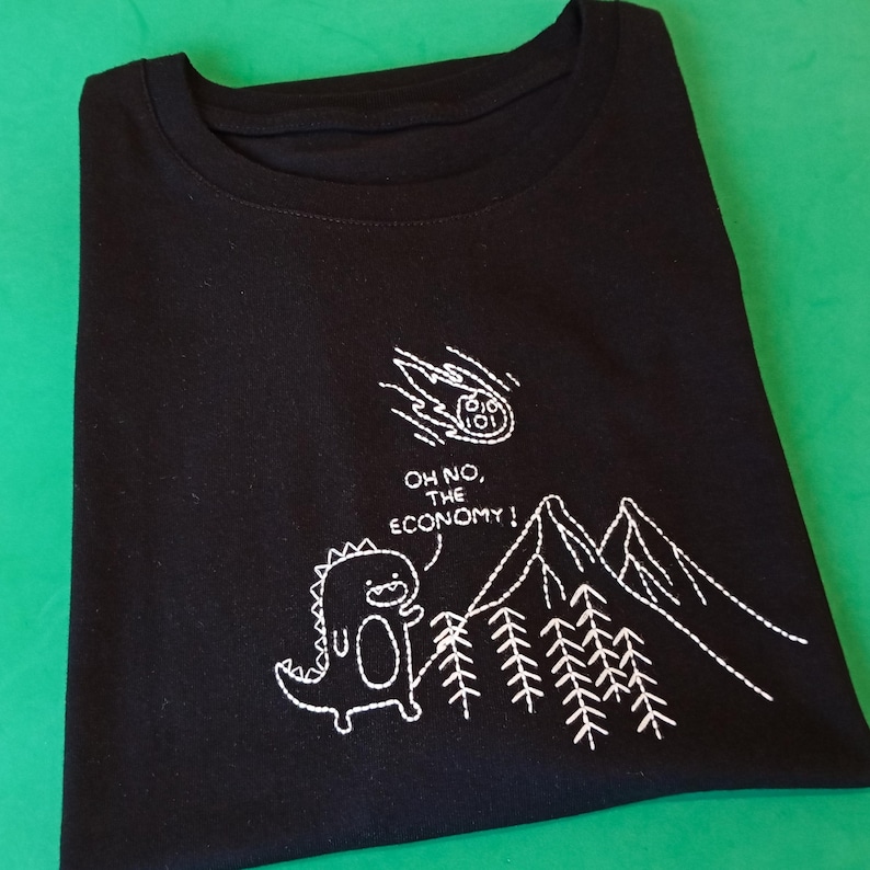 May include: Black t-shirt with white embroidered design of a dinosaur looking up at a falling comet labeled "The Economy". The text "Oh no, the economy!" is embroidered below the comet.