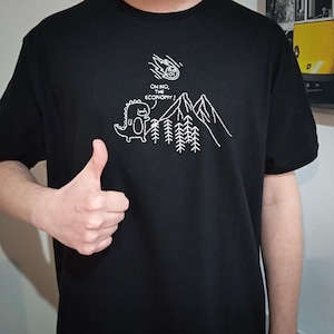 May include: Black t-shirt with a white embroidered design of a dinosaur standing in front of a mountain range with trees. The dinosaur is looking up at a falling meteor with the text "Oh no, the economy!" above it.