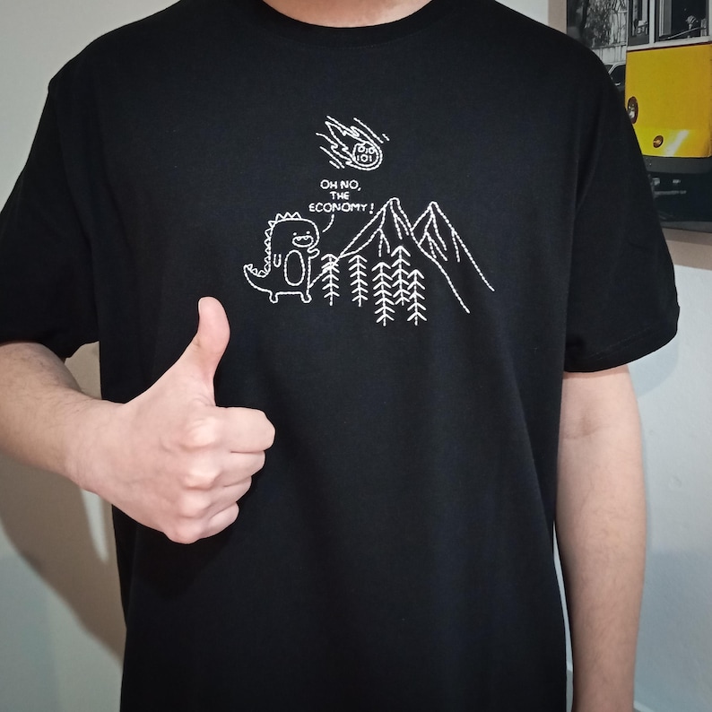 May include: Black t-shirt with a white embroidered design of a dinosaur standing in front of a mountain range with trees. The dinosaur is looking up at a falling meteor with the text "Oh no, the economy!" above it.