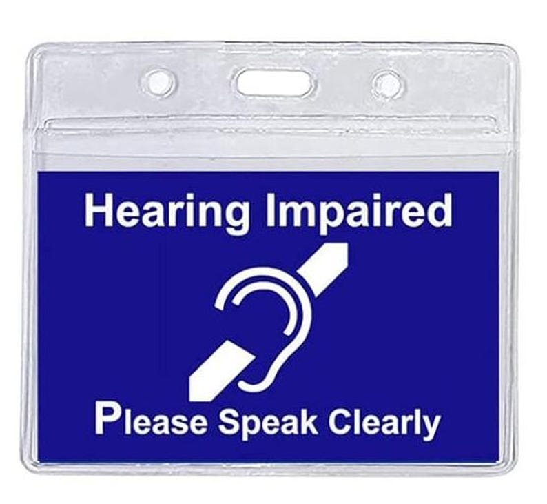 Puede incluir: Un portagafete de pl&aacute;stico transparente con un inserto azul que dice "Hearing Impaired" y un s&iacute;mbolo blanco de una oreja tachada. El texto "Please Speak Clearly" tambi&eacute;n est&aacute; impreso en blanco en el inserto azul.