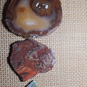 May include: Three agate slices, one with brown and white banding, one with red and brown banding, and one with green and pink banding. The slices are on a woven surface.