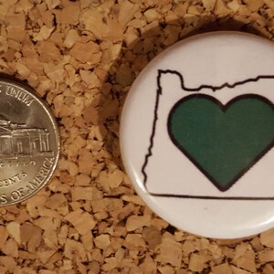 May include: A white button with a black outline of the state of Oregon and a green heart in the center.