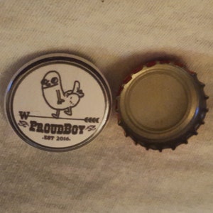 Pode incluir: Uma tampa de garrafa prateada com fundo branco e uma ilustração em preto e branco de um personagem de desenho animado. O texto "Proudboy" é impresso em preto com o texto "Est. 2016" abaixo.