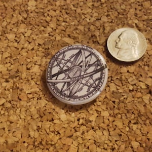 May include: A white button with a black and white image of a celestial sphere with lines and circles. The button is on a brown corkboard.