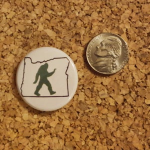 May include: A white button featuring a green silhouette of Bigfoot walking within the outline of the state of Oregon.
