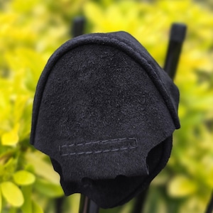 Könnte beinhalten: Eine schwarze Golfschlägerhaube aus Wildleder. Die Hülle hat eine dunkle, einfarbige Farbe mit einem genähten Detail. Der Hintergrund ist eine verschwommene Ansicht von grünem Laub.