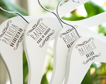 May include: White wooden hangers with black lettering. The hangers are personalized with names and titles such as "Bride", "Maid of Honor", and "Bridesmaid".
