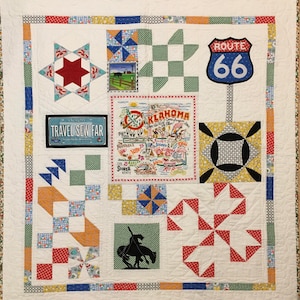 Op de afbeelding: Een wit quilt met een verscheidenheid aan kleurrijke patchwork-vierkanten. De vierkanten hebben verschillende ontwerpen, waaronder sterren, windmolens en een kaart van Oklahoma. Het quilt bevat ook een bord met de tekst "Route 66" en een bord met de tekst "Travel Sew Far".