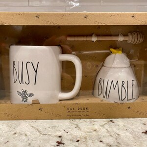 Può includere: Un set di tazza e vasetto di miele in ceramica bianca con un design a forma di ape. La tazza dice "Busy" e il vasetto di miele dice "Bumble". Il set viene fornito in una scatola marrone con un motivo a forma di ape.