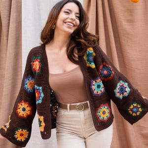 Peut inclure: Une femme portant un cardigan en crochet marron avec des accents floraux colorés. Le cardigan a un col en V et des manches longues. Elle porte un pantalon blanc et sourit.