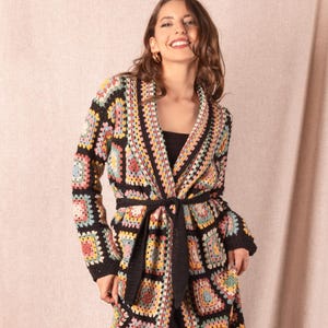 Peut inclure: Longue veste en crochet ouverte, avec un motif patchwork. La veste présente une base noire avec des carrés colorés dans les tons de jaune, rose, vert et bleu. Elle a des manches longues et une ceinture noire assortie. La veste mesure environ 140 cm de long.
