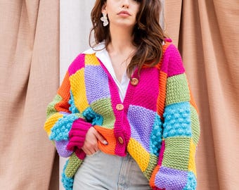 Może przedstawiać: Kobieta w kolorowym kardiganie w stylu patchwork z zapięciem na guziki. Kardigan wykonany jest z kwadratów z przędzy w różnych kolorach, w tym różowym, fioletowym, zielonym, żółtym i pomarańczowym. Kardigan ma luźny krój i jest noszony na białej koszuli. Kobieta ma na sobie niebieskie dżinsy.
