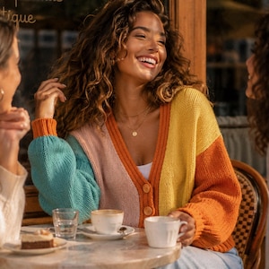 Peut inclure: Un cardigan multicolore avec des panneaux turquoise, roses, oranges et jaunes. Le cardigan est boutonné et porté avec un haut blanc et un jean bleu clair. Une femme sourit à une table de café avec du café et une pâtisserie.