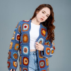Könnte beinhalten: Ein blauer Häkelcardigan mit bunten geometrischen Mustern. Der Cardigan ist vorne offen und hat lange Ärmel.