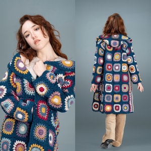 May include: A blue crocheted cardigan with a hood, featuring a colourful geometric pattern of squares and circles. The cardigan is being worn by a person with long brown hair.