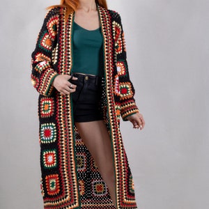 K&ouml;nnte beinhalten: Ein langer, schwarzer H&auml;kelcardigan mit bunten geometrischen Mustern. Der Cardigan hat einen tiefen V-Ausschnitt und ist vorne offen. Das Model tr&auml;gt den Cardigan &uuml;ber einem gr&uuml;nen Tanktop und schwarzen Shorts.