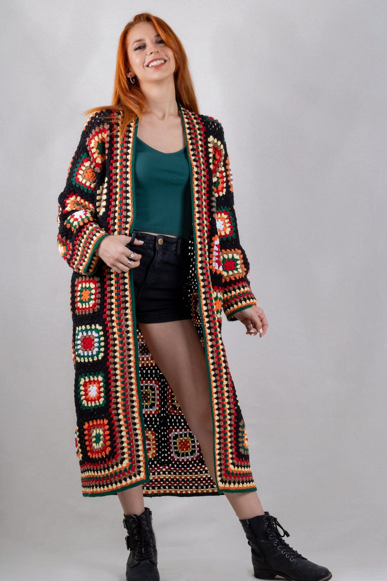 K&ouml;nnte beinhalten: Ein langer, schwarzer H&auml;kelcardigan mit bunten geometrischen Mustern. Der Cardigan hat einen tiefen V-Ausschnitt und ist vorne offen. Das Model tr&auml;gt den Cardigan &uuml;ber einem gr&uuml;nen Tanktop und schwarzen Shorts.