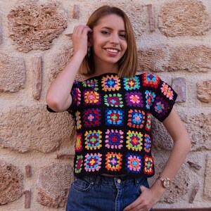 Puede incluir: Una blusa de ganchillo de manga corta construida a partir de una cuadr&iacute;cula de cuadrados de la abuela multicolores con tonos vibrantes de rosa, azul, amarillo, verde y morado, todos bordeados con hilo negro, combinada con jeans de mezclilla azul.