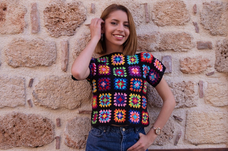 Puede incluir: Una blusa de ganchillo de manga corta construida a partir de una cuadr&iacute;cula de cuadrados de la abuela multicolores con tonos vibrantes de rosa, azul, amarillo, verde y morado, todos bordeados con hilo negro, combinada con jeans de mezclilla azul.