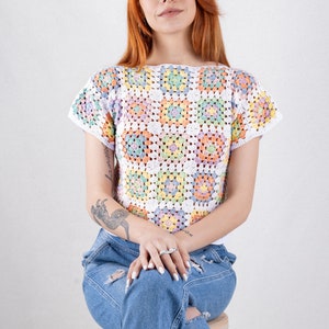 Puede incluir: Un top de ganchillo de manga corta construido a partir de una serie de motivos cuadrados en tonos pastel de amarillo, rosa, azul y verde, todos bordeados con hilo blanco. La prenda presenta un escote ancho y un corte corto, combinado con vaqueros azules.