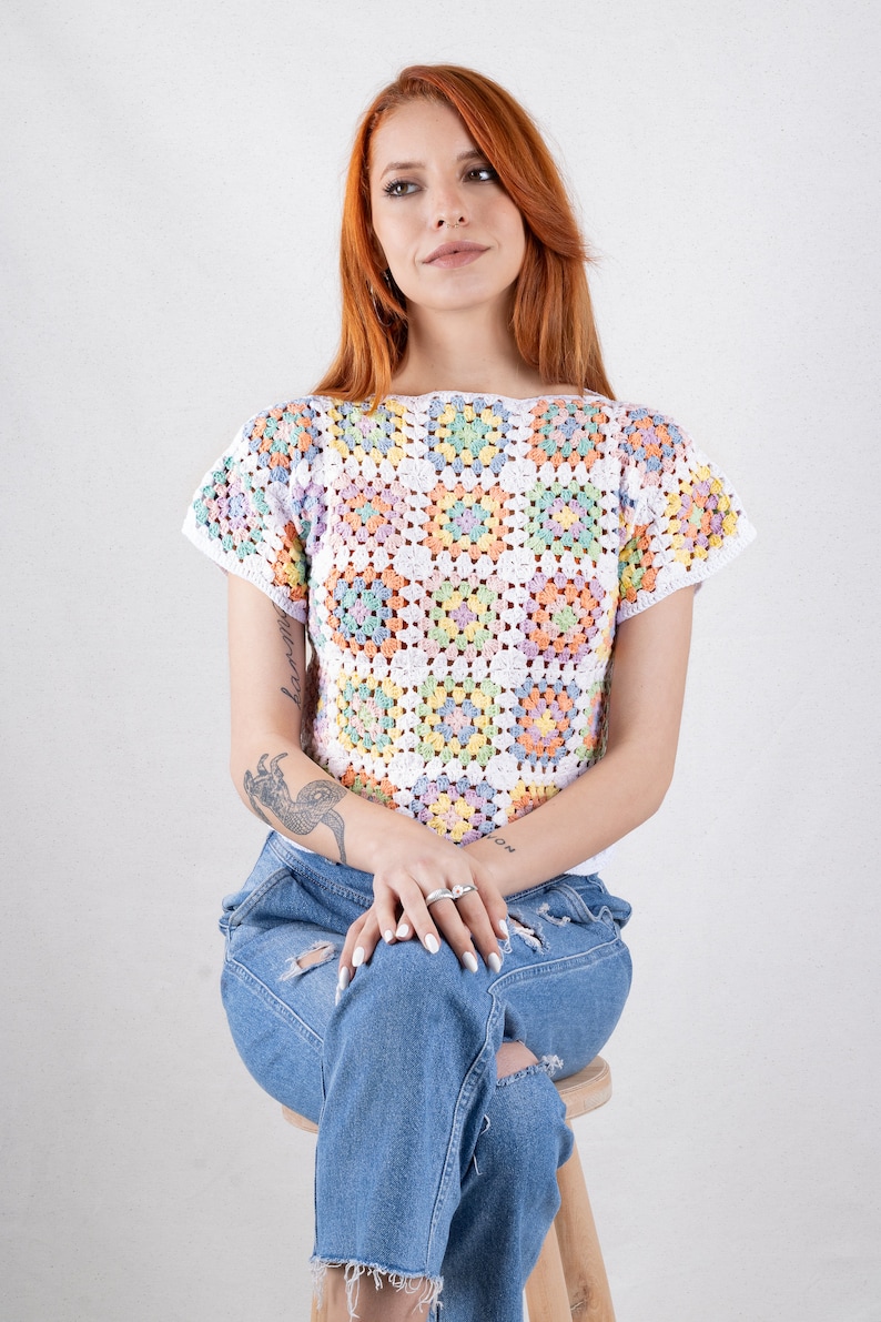 Puede incluir: Un top de ganchillo de manga corta construido a partir de una serie de motivos cuadrados en tonos pastel de amarillo, rosa, azul y verde, todos bordeados con hilo blanco. La prenda presenta un escote ancho y un corte corto, combinado con vaqueros azules.